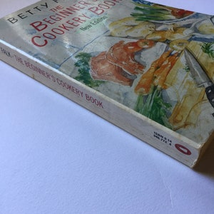 May include: A white cookbook with a red and green illustration of vegetables and seafood on the cover. The title is "The Beginner's Cookery Book" by Betty Falk. The book is a new edition.