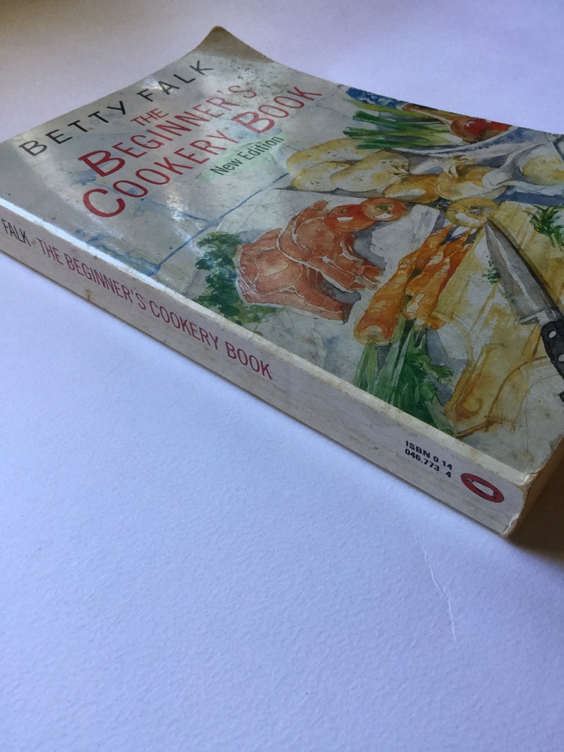 May include: A white cookbook with a red and green illustration of vegetables and seafood on the cover. The title is "The Beginner's Cookery Book" by Betty Falk. The book is a new edition.