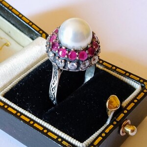 Può includere: Anello d'argento con una grande perla bianca rotonda al centro, circondata da un'aureola di piccole pietre preziose rosse e trasparenti. L'anello è presentato in un portagioie nero con finiture dorate.
