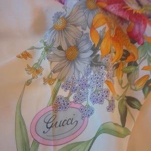 May include: A white silk scarf with a floral print featuring lilies, daisies, and other flowers. The scarf has a pink oval label with the word "Gucci" written in cursive.