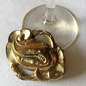 Può includere: Un oggetto metallico dorato e ornato con un design floreale, forse una spilla o un elemento decorativo. L'oggetto è posizionato vicino alla base di un gambo di vetro trasparente. Lo sfondo è una superficie bianca.