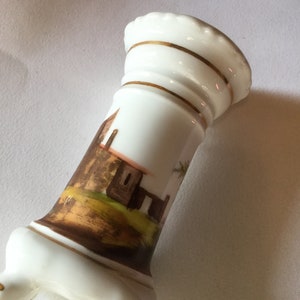 May include: A white porcelain vase with a gold trim and a painted scene of a building and trees. The vase has three legs and is approximately 6 inches tall.