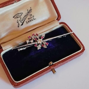 May include: A vintage brooch in a red velvet box. The brooch is made of silver and features a starburst design with red and white stones. The box is labelled "Watchmaker and Jeweller J. Carmichael 28 Main St. LARGS."