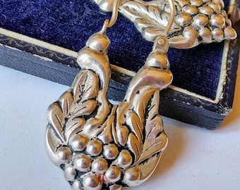 May include: A pair of silver earrings with a grape cluster design. The earrings are detailed with leaves and have a hoop style.
