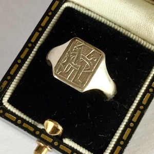 Può includere: Un anello a sigillo in argento con una superficie quadrata che presenta un monogramma inciso. L'anello è esposto in una scatola per anelli in velluto nero con una chiusura dorata.