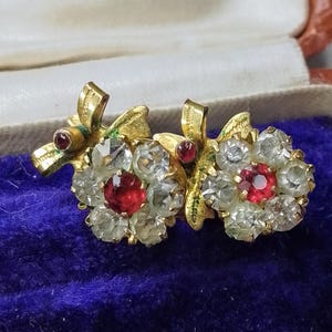 May include: A pair of gold tone earrings with a floral design. Each earring features a cluster of clear rhinestones and a single red rhinestone in the center.