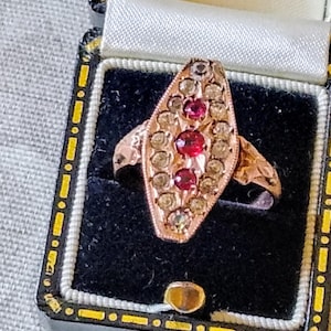 Puede incluir: Un anillo de oro vintage con una cara en forma de marquesa, engastado con pequeñas piedras preciosas claras y rojas. El anillo se exhibe en un joyero negro con ribete dorado e interior de satén blanco. El diseño del anillo presenta detalles intrincados.