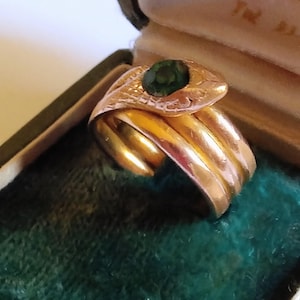Könnte beinhalten: Ein goldfarbener Ring in Form einer gewundenen Schlange mit strukturiertem Kopf und glatter, mehrlagiger Ringschiene. Der Schlangenkopf ist mit einem einzelnen, runden, dunkelgrünen Edelstein in der Mitte besetzt. Der Ring liegt in einer dunkelgrünen Samtschachtel.
