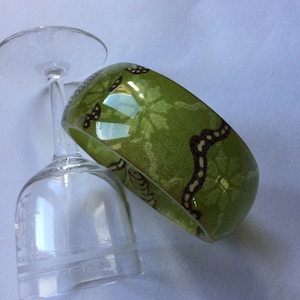 Könnte beinhalten: Ein grünes Armband mit braunem Blumenmuster. Das Armband ist aus Kunststoff und hat eine glänzende Oberfläche.