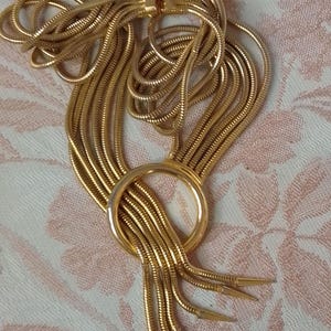 Op de afbeelding: Een meerrijige gouden ketting met een grote ronde hanger en puntige uiteinden.