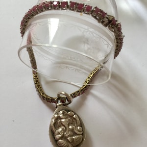 Può includere: Una collana a catena d'argento con un ciondolo che presenta una rappresentazione dettagliata della divinità indù Ganesha. La catena è adornata da pietre preziose rosa.