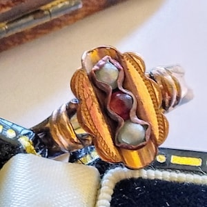 Puede incluir: Primer plano de un anillo vintage dorado con un diseño floral. El anillo presenta tres piedras incrustadas: una verde claro, una roja y una gris claro. El anillo está sobre un joyero.