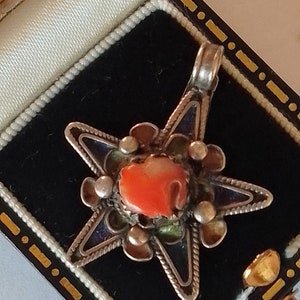 Puede incluir: Un colgante de plata en forma de estrella con una piedra de coral en el centro. El colgante está decorado con esmalte azul y cuentas de plata.