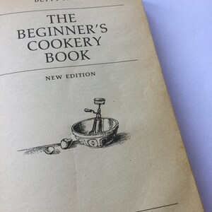 May include: The cover of a vintage cookbook titled "The Beginner's Cookery Book" by Betty Falk. The cover features a black and white illustration of a mixing bowl with a hand-crank mixer and a broken egg.