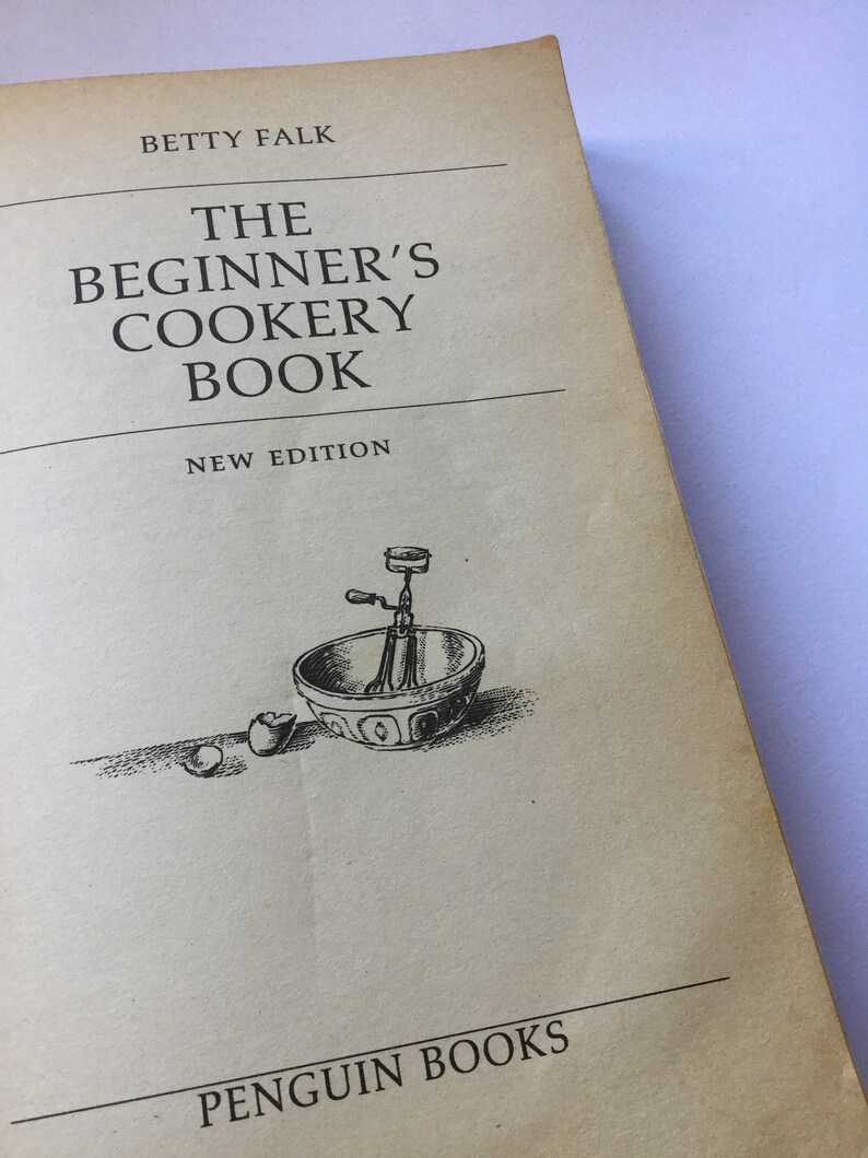 May include: The cover of a vintage cookbook titled "The Beginner's Cookery Book" by Betty Falk. The cover features a black and white illustration of a mixing bowl with a hand-crank mixer and a broken egg.