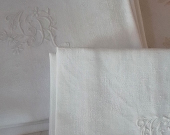 May include: A stack of white linen napkins with embroidered initials and floral designs. The napkins are folded and stacked on a patterned surface.