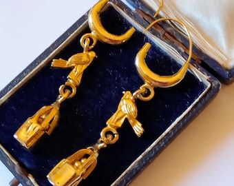 Puede incluir: Un par de pendientes dorados con un pájaro posado sobre un dije en forma de jaula. Los pendientes tienen un diseño de aro y se exhiben en un joyero azul vintage. Los pendientes probablemente son de metal dorado.