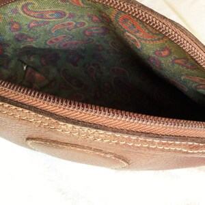 May include: A brown leather zippered pouch with a paisley patterned lining. The pouch is closed and the zipper is visible.