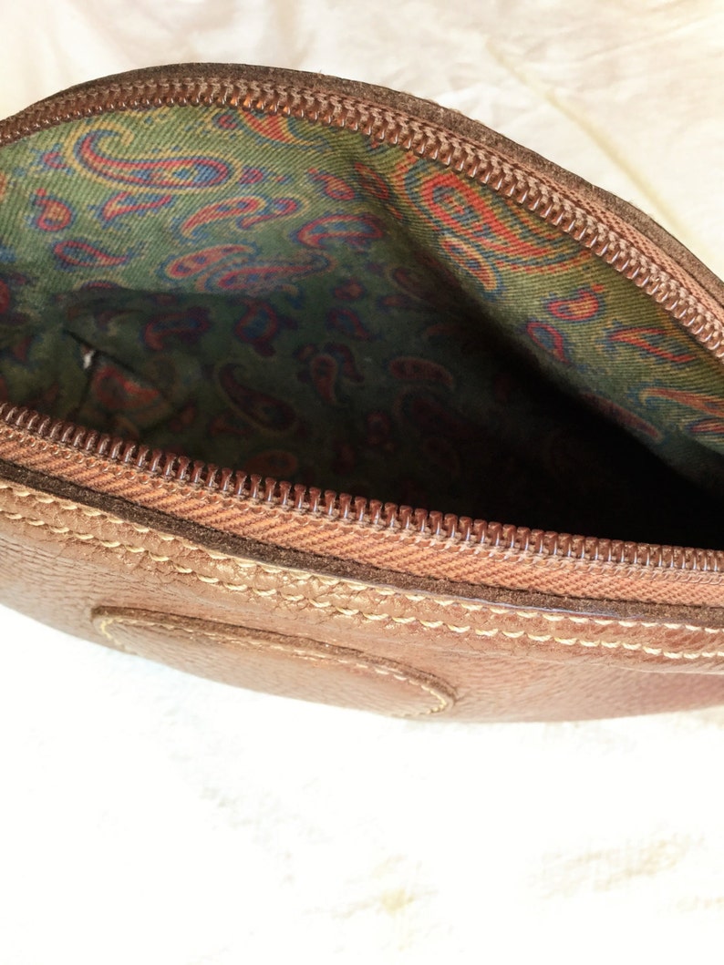 May include: A brown leather zippered pouch with a paisley patterned lining. The pouch is closed and the zipper is visible.