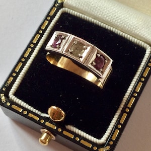 Peut inclure: Une bague en or avec trois pierres précieuses, deux rouges et une verte, serties dans un bandeau argenté. La bague est présentée dans un écrin noir avec un intérieur blanc et des détails dorés. La bande de la bague est en or.