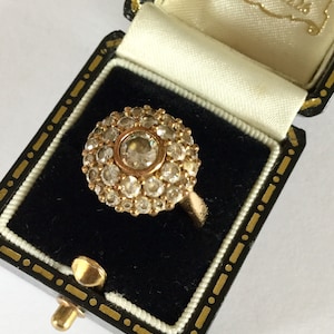 Può includere: Un anello d'oro con una grande pietra rotonda centrale circondata da pietre più piccole. L'anello è in una scatola per anelli nera con un interno bianco.