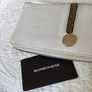 May include: White leather wallet with a brown leather accent and a silver metal logo that says "Borbonese". The wallet is closed and lying on a white textured surface. A black card with the word "Borbonese" printed in white is lying on the surface.