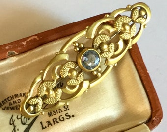 May include: A gold-toned brooch with a delicate, intricate design featuring a light blue gemstone in the center. The brooch is in a small, brown velvet box with the text "J.W. Main St. LARGS." on the inside.