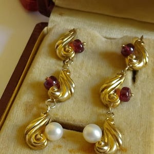 Può includere: Un paio di orecchini pendenti color oro con un design a spirale. Ogni orecchino presenta una piccola gemma rossa rotonda, una perla bianca più grande ed elementi dorati curvi. Gli orecchini sono esposti in un portagioie vintage.
