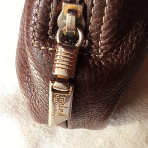 May include: Close-up of a brown leather zipper pull with a gold metal zipper slider and a gold metal tag that reads "redwall Made in Italy".