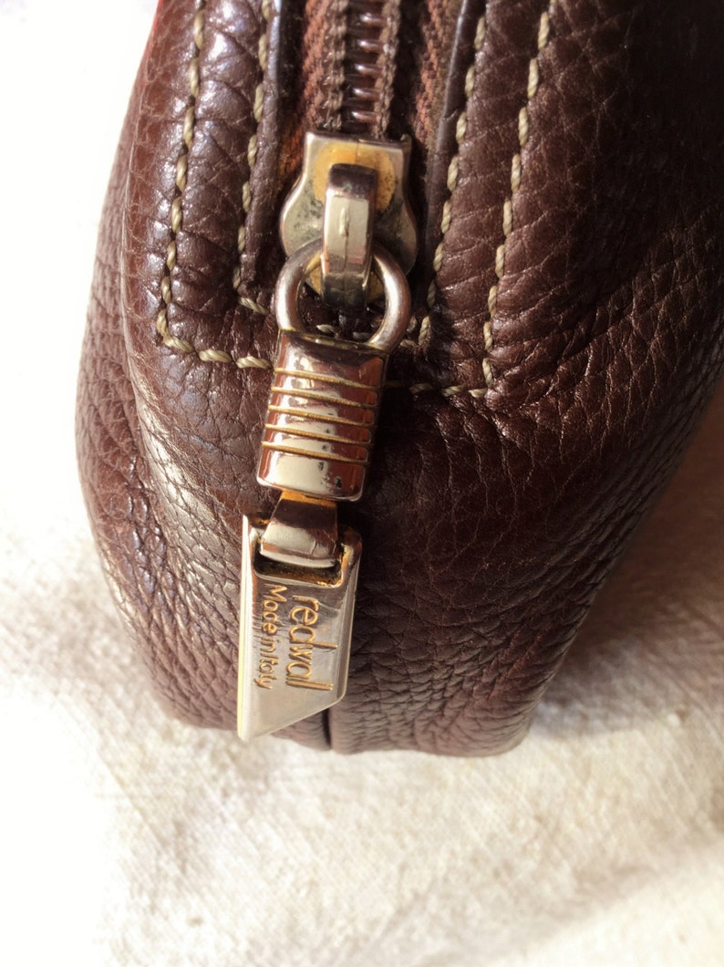 May include: Close-up of a brown leather zipper pull with a gold metal zipper slider and a gold metal tag that reads "redwall Made in Italy".