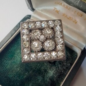 Può includere: Un anello d'argento con una montatura quadrata che presenta un gruppo di piccole pietre trasparenti. L'anello è in una scatola di velluto verde.