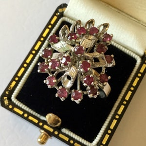 Può includere: Un anello color argento con un gruppo di piccole gemme rosse rotonde. L'anello ha un design floreale ed è presentato in un portagioie nero con dettagli dorati.