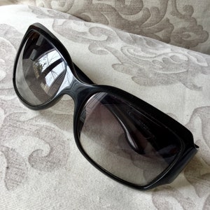 Puede incluir: Gafas de sol negras con montura rectangular y lentes oscuras. Las gafas de sol están apoyadas sobre una superficie de tela estampada.