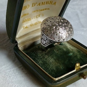Può includere: Un anello d'argento con un designo circolare testurizzato. L'anello è in una scatola di velluto verde con un rivestimento in raso bianco. La scatola ha una chiusura dorata e il testo "D'AMBRA SIMO D'AMBRA GIOELLERIA OROLOGERIA MARINO (CT) Tel. 93-20-24" è stampato sul rivestimento.
