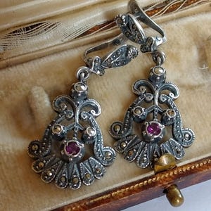 Può includere: Un paio di orecchini antichi in argento con un design floreale, incastonati con piccole pietre di marcasite e una pietra centrale color rubino. Gli orecchini sono esposti in un portagioie vintage.