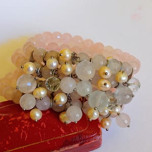 May include: A multi-strand bracelet featuring rose quartz beads and dangling charms with pearls and clear quartz. The bracelet is resting on a red jewelry box. The beads are in shades of pink, cream, and clear.