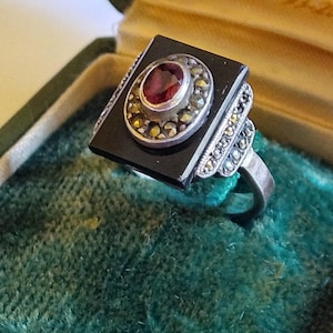 Può includere: Anello vintage in argento con una pietra rettangolare di onice nera, un rubino ovale centrale e dettagli in marcasite. L'anello è incastonato in una fascia d'argento e presentato in un portagioie di velluto verde.
