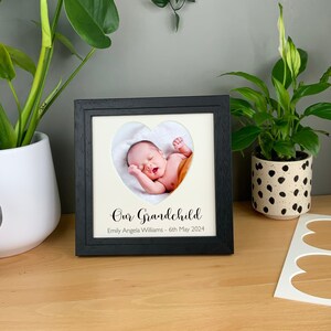 May include: Black picture frame with a white mat and a heart-shaped cutout. The cutout shows a photo of a baby sleeping. The text "Our Grandchild" is printed below the photo, followed by "Emily Angela Williams - 6th May 2024".