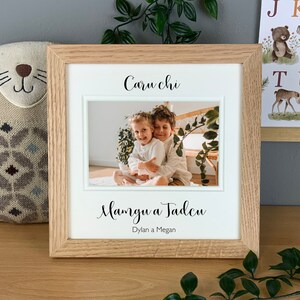 May include: A wooden picture frame with a white mat and a photo of two children inside. The frame has the text "Caru chi" and "Mamgu a Tadcu" written in cursive on it. The text "Dylan a Megan" is written in a smaller font below the cursive text.
