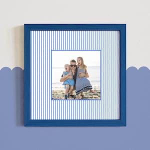 May include: A square blue picture frame with a family photo. The frame has a blue and white striped background. The photo shows a family of three on a beach. The frame is mounted on a blue wall with a scalloped design.