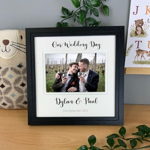 May include: Black framed photo with a white mat. The photo shows two people smiling and holding wine glasses. The text "Our Wedding Day" is at the top of the mat. The text "Dylan & Paul" is below the photo. The date "23rd September 2023" is below the names.