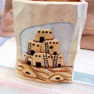May include: A white ceramic vase with a painted scene of a pueblo-style building with ladders. The vase has a brown rim and a textured surface.