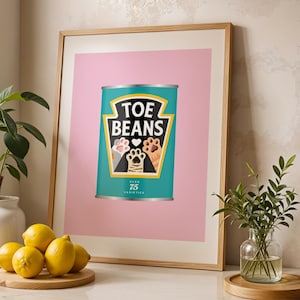 May include: Framed print with a teal can design, labelled "TOE BEANS" with cat paw illustrations. The print is set against a pink background and framed in light wood. The image is displayed on a countertop with lemons and plants.
