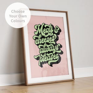 May include: A framed wall art print featuring the text Meats & cheeses always pleases in a stylised green font with a black drop shadow, set against a solid light pink background. The frame is a medium-toned wood finish.