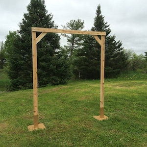 May include: A wooden wedding archway with a simple design, standing on a grassy lawn. The archway is made of natural wood and has a rustic look.
