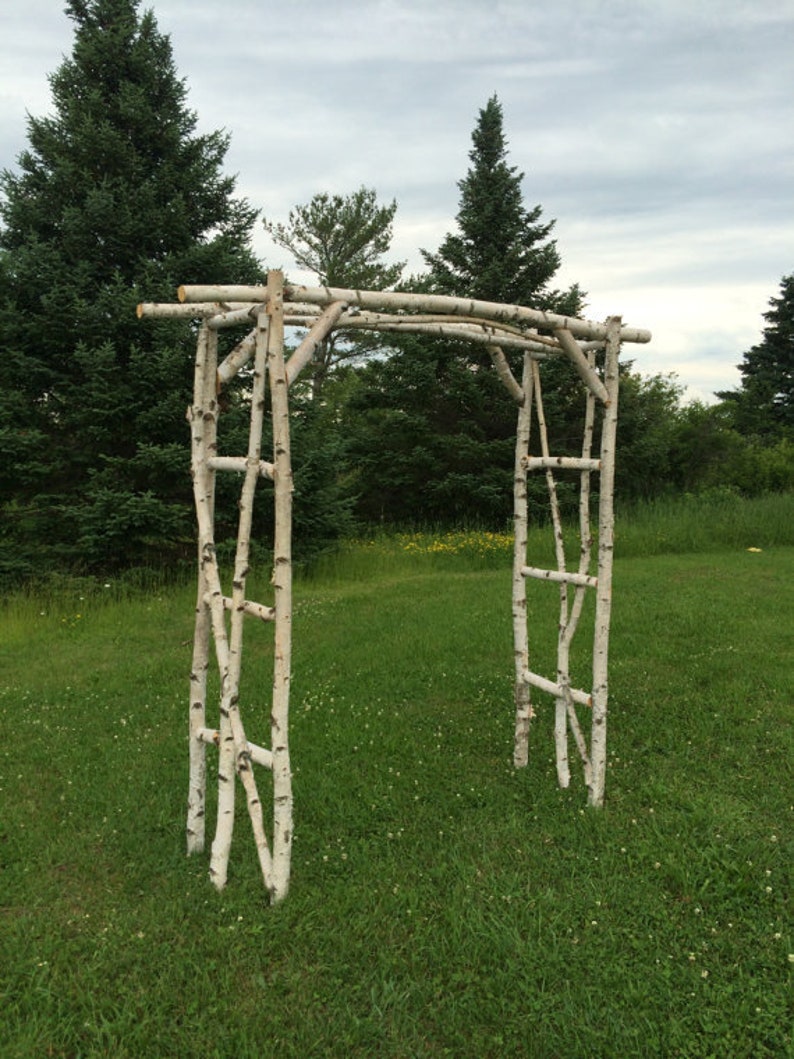 May include: A white birch branch archway for a wedding ceremony or other outdoor event. The archway is made of natural birch branches and is standing on a grassy lawn.