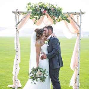 May include: A bride and groom kiss under a birchwood archway decorated with greenery and pink flowers. The archway is draped with sheer peach fabric. The bride is wearing a white strapless wedding dress and a veil. The groom is wearing a gray suit. The couple is standing on a green grassy lawn.