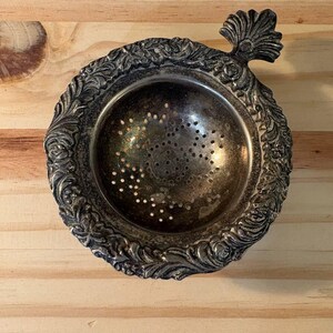 May include: A silver-toned metal tea strainer with a circular bowl featuring a perforated base. The rim is decorated with an embossed floral and leaf pattern, and a small, fan-shaped handle extends from one side of the circular frame.