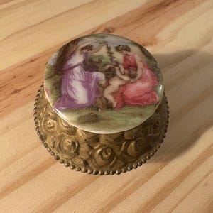 Könnte beinhalten: Kleine, antike Schmuckdose mit detaillierter, verzierter Messingbasis. Der Deckel zeigt eine bemalte Szene von Figuren in Rosa-, Lila- und Grüntönen. Die Dose ist rund und steht auf einer Holzoberfläche.