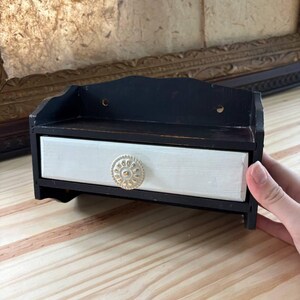 May include: A small wooden wall-mounted shelf featuring a black frame and a single white drawer. The drawer is accented with a round, textured, metallic-colored knob. The unit has two circular holes on the back panel for mounting purposes.